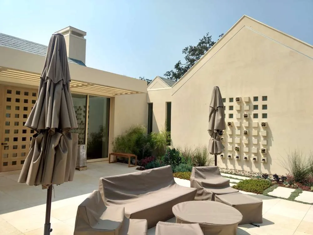 Outdoor patio with beige stucco walls, covered furniture, and potted plants under a sunny sky.