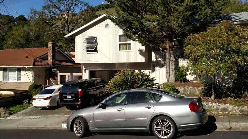 Street view of a two-story house with vertical siding, trees, and parked cars.