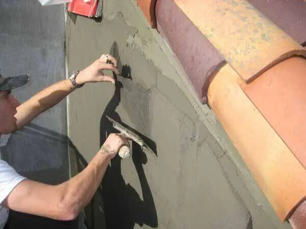Worker applying stucco over mesh on exterior wall near terracotta roof tiles.