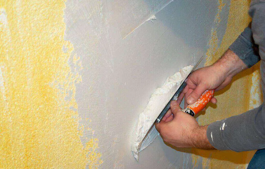Person applying white plaster to a yellow wall with a trowel.