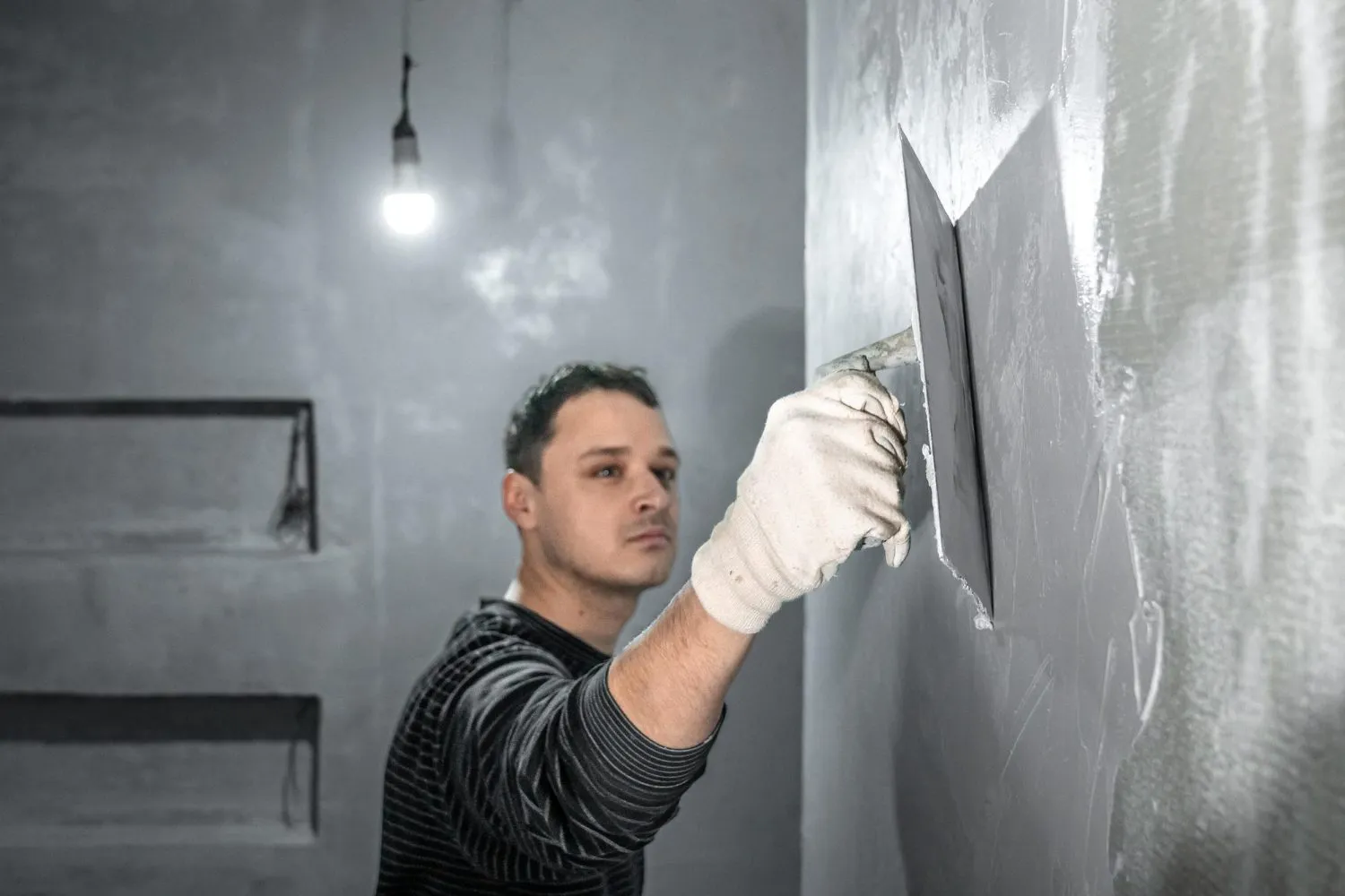 Person applying plaster to a wall with a trowel in a dimly lit room.